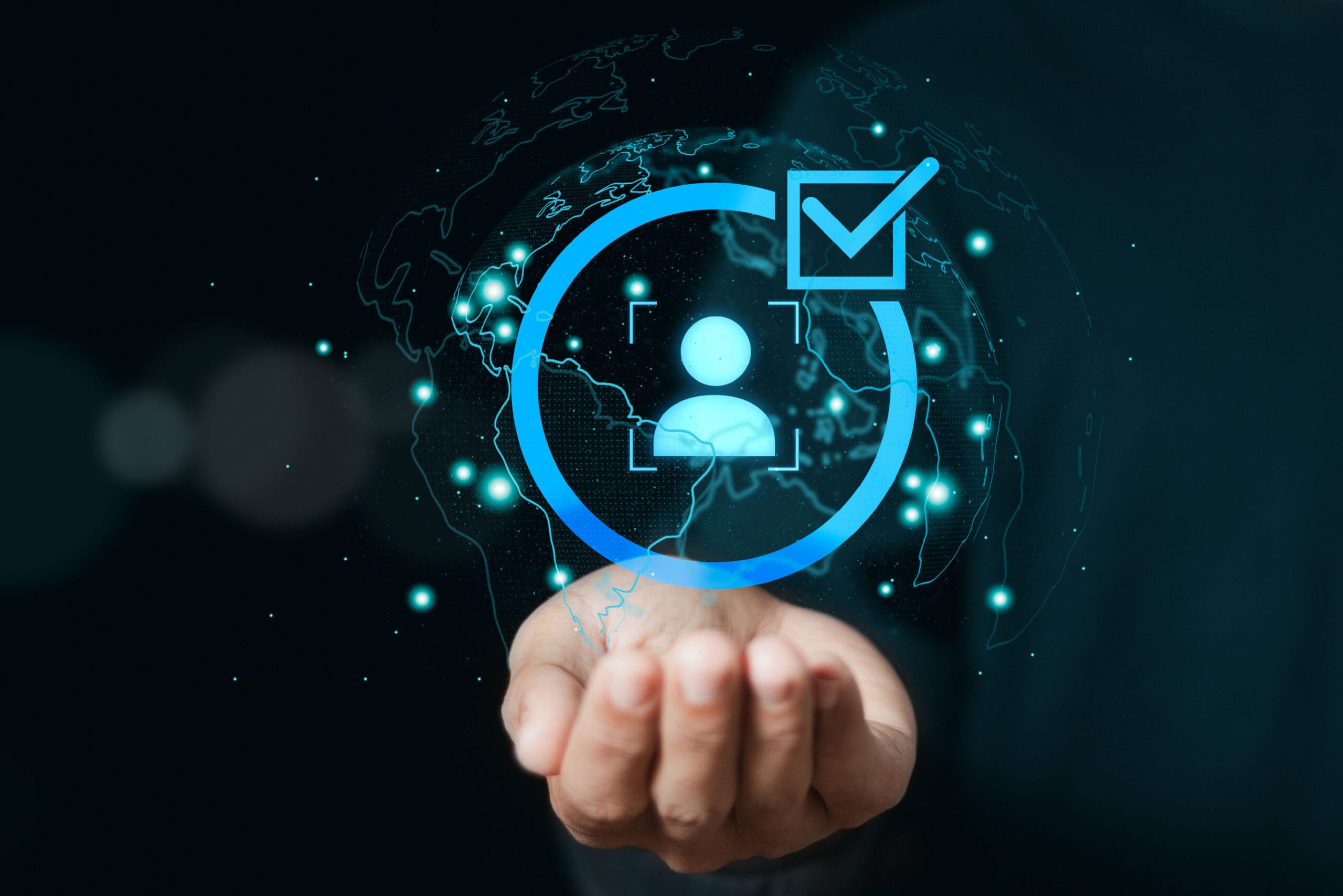 A hand appearing to hold or project a glowing blue circular hologram containing a user profile icon and a checkmark symbol. The image has a dark background with bokeh effects and small particle elements, suggesting a digital verification or authentication process.