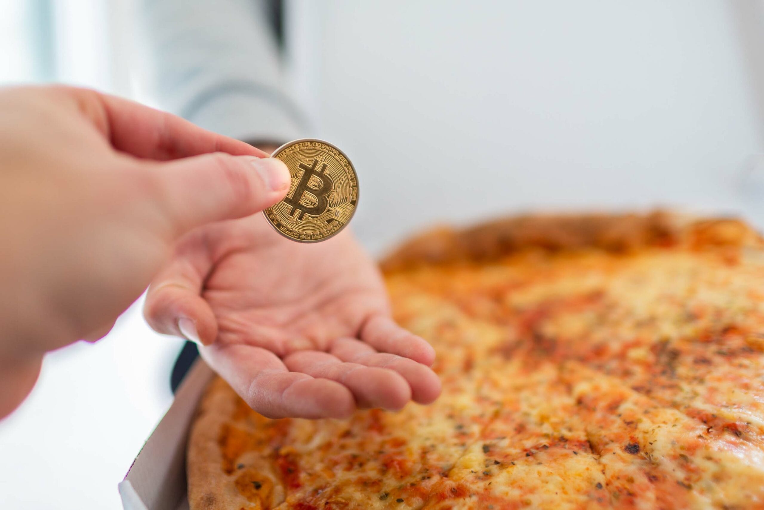 A hand holding a physical Bitcoin coin over a freshly baked pizza, creating a visual reference to "Bitcoin Pizza Day" - when the first real-world Bitcoin transaction was used to purchase two pizzas. The pizza appears to be a cheese pizza and is in soft focus in the background.