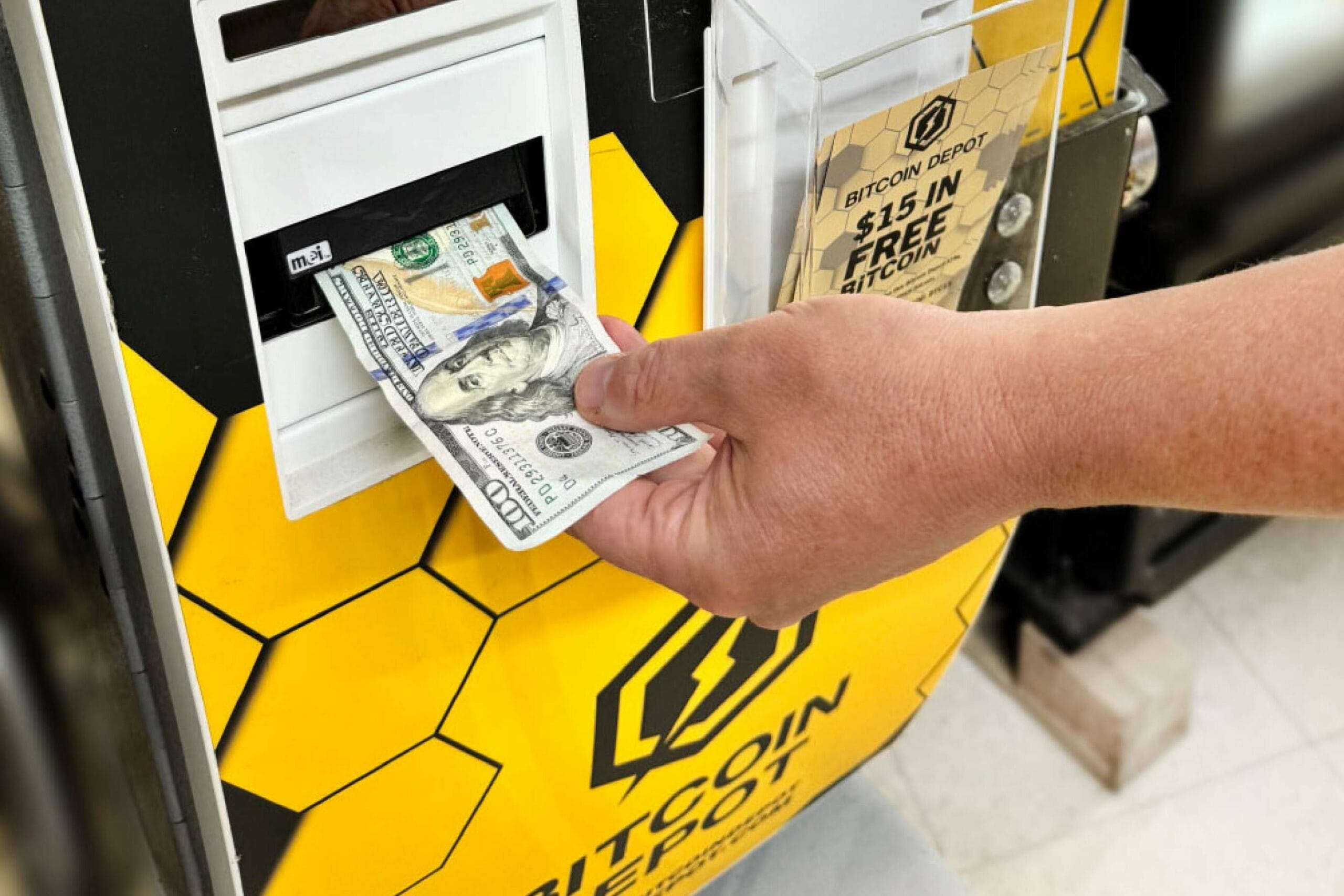 A hand inserts a $100 bill into a Bitcoin ATM. The machine is yellow and black with a honeycomb pattern. A sign on the ATM reads 'BITCOIN DEPOT $15 IN FREE BITCOIN', indicating a promotional offer.