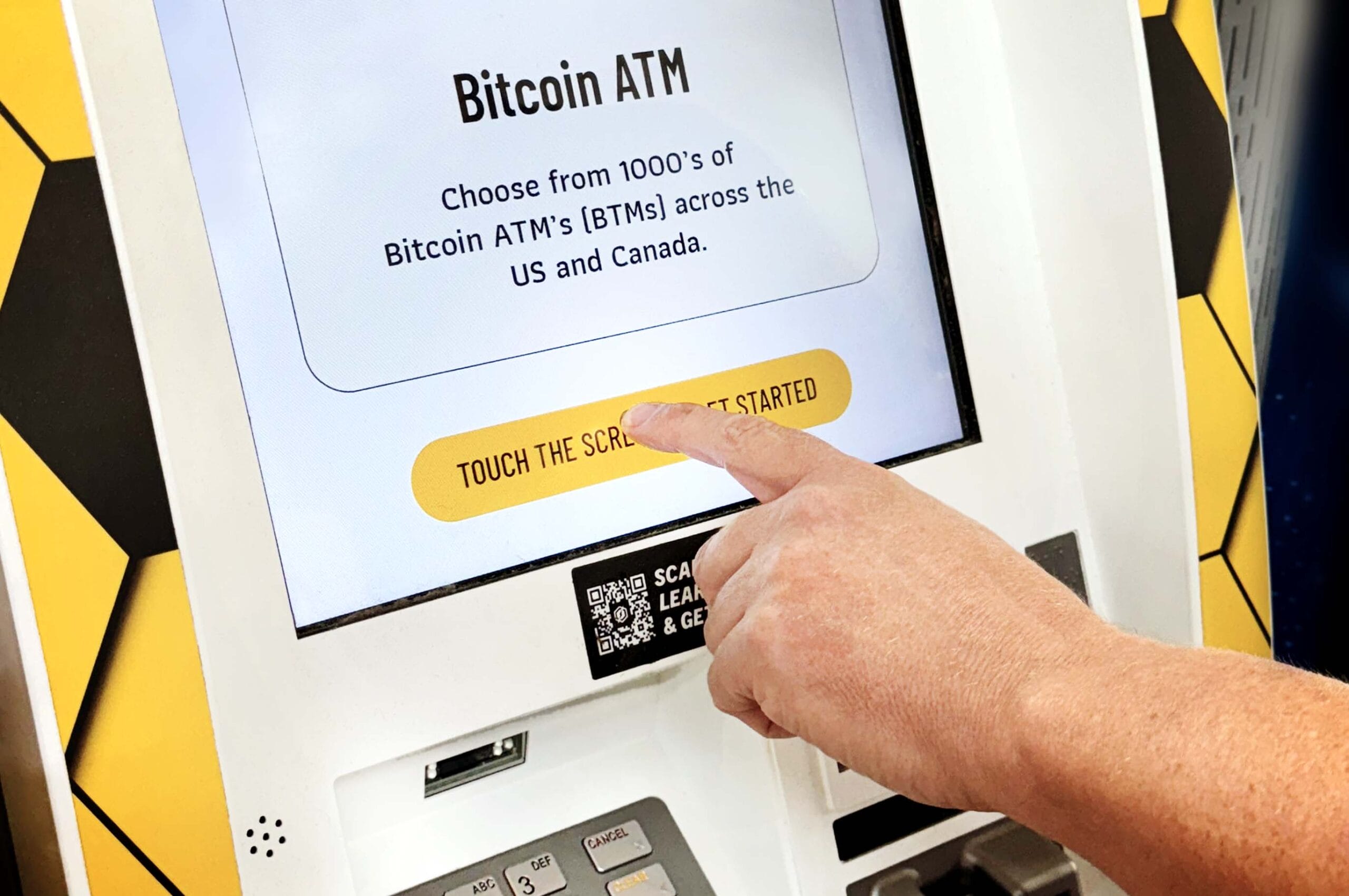 A close-up of a Bitcoin ATM screen showing a welcome message and a yellow 'TOUCH THE SCREEN TO GET STARTED' button. A hand is reaching out to touch the screen. The ATM display indicates it's part of a network of Bitcoin ATMs across the US and Canada.