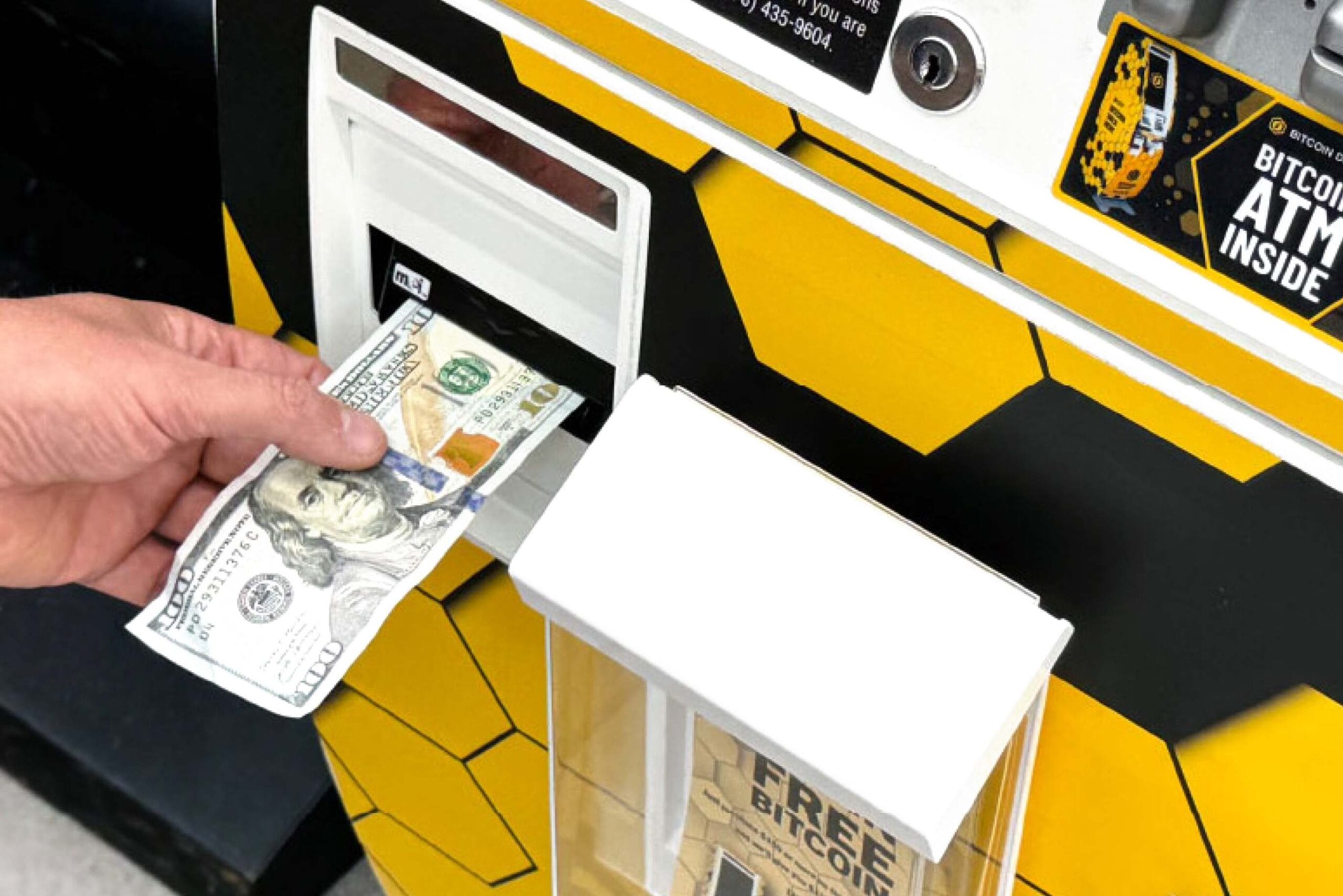 A hand inserting a $100 bill into a Bitcoin ATM with a yellow and black geometric design. The ATM displays "BITCOIN ATM INSIDE" signage in the top right corner, and there appears to be a bill acceptor slot where the cash is being inserted.