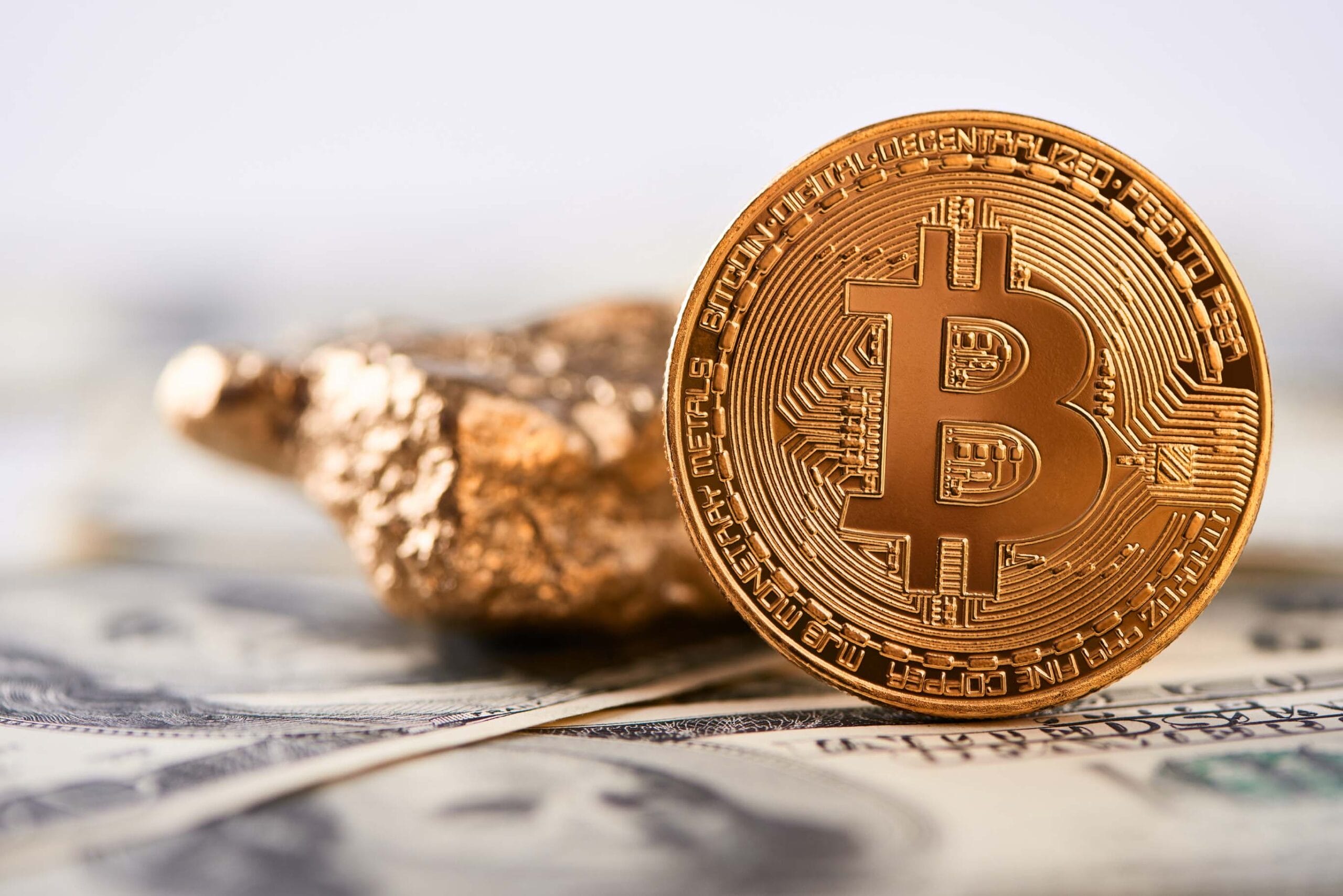 A close-up of a physical Bitcoin coin standing upright on U.S. dollar bills, with a gold nugget in the background. The image symbolizes the comparison between Bitcoin and traditional assets like gold and fiat currency.