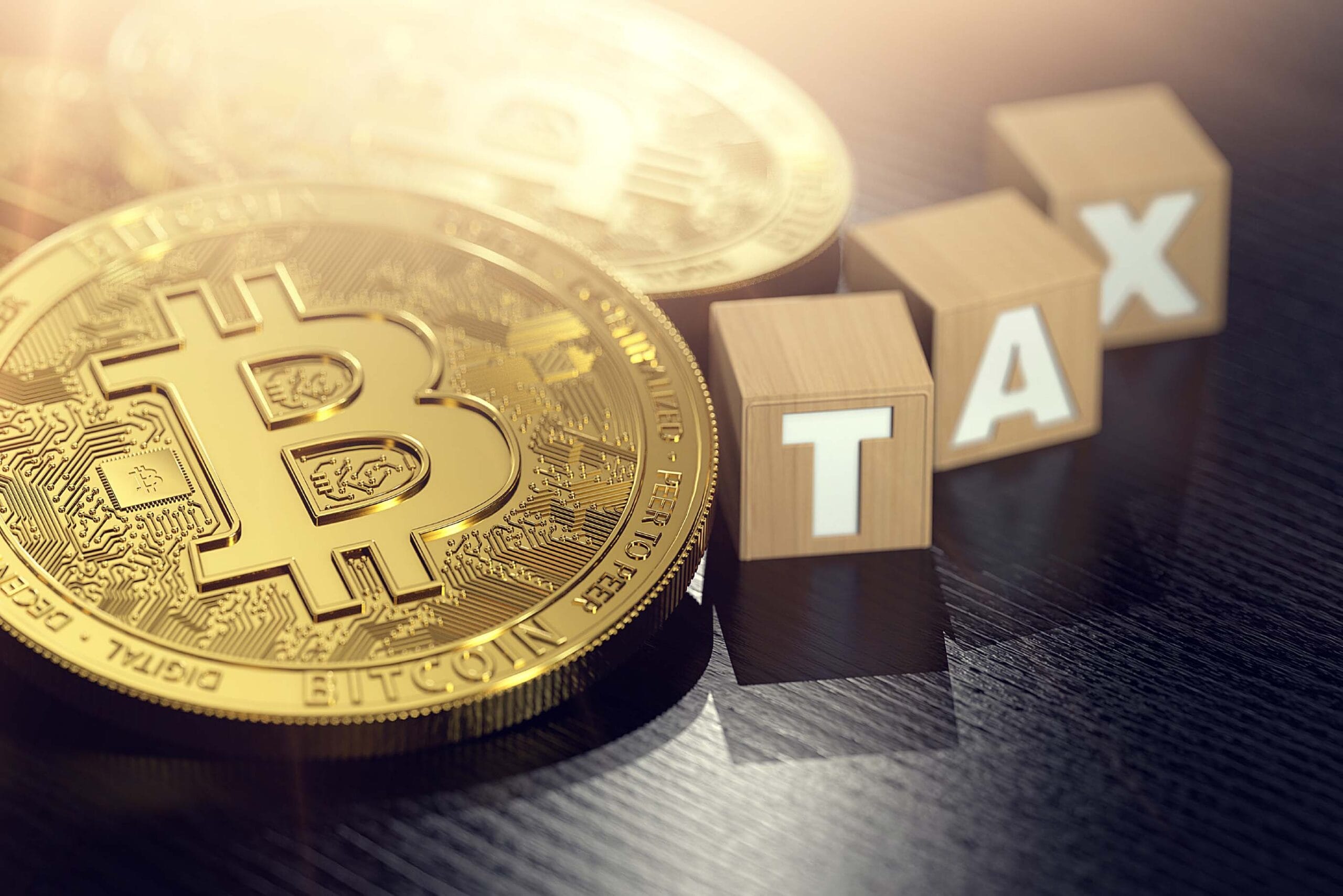 Close-up of shiny gold Bitcoin coins resting on a dark reflective surface, accompanied by wooden blocks spelling out the word 'TAX' in white capital letters. The image represents cryptocurrency taxation and financial regulations, with a warm light effect adding emphasis.