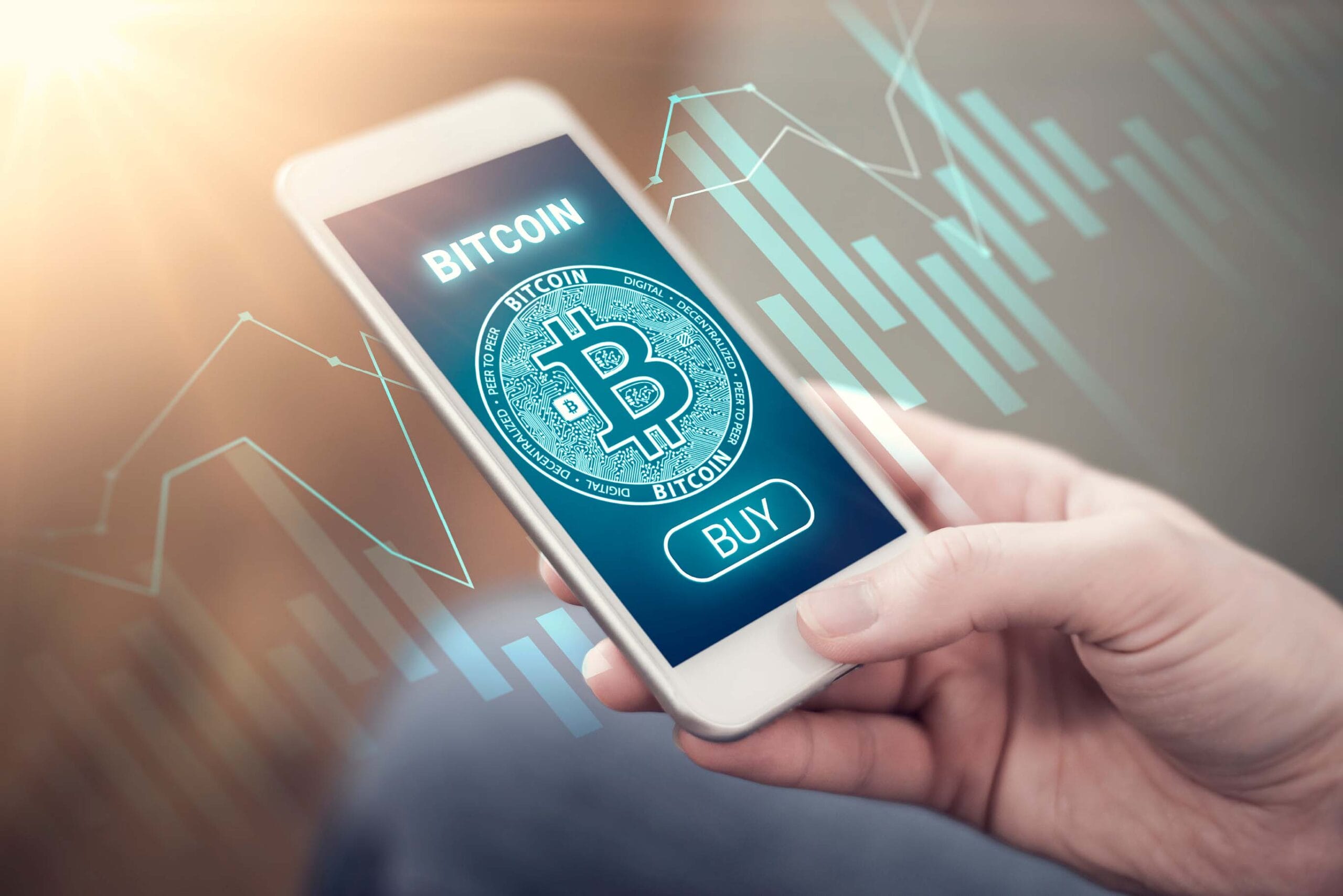A hand holding a smartphone displaying a Bitcoin trading app screen with a glowing Bitcoin logo, "BITCOIN" text, and a "BUY" button in blue and white colors. Overlaid on the background are light blue digital graphs and charts suggesting market data or price movements.