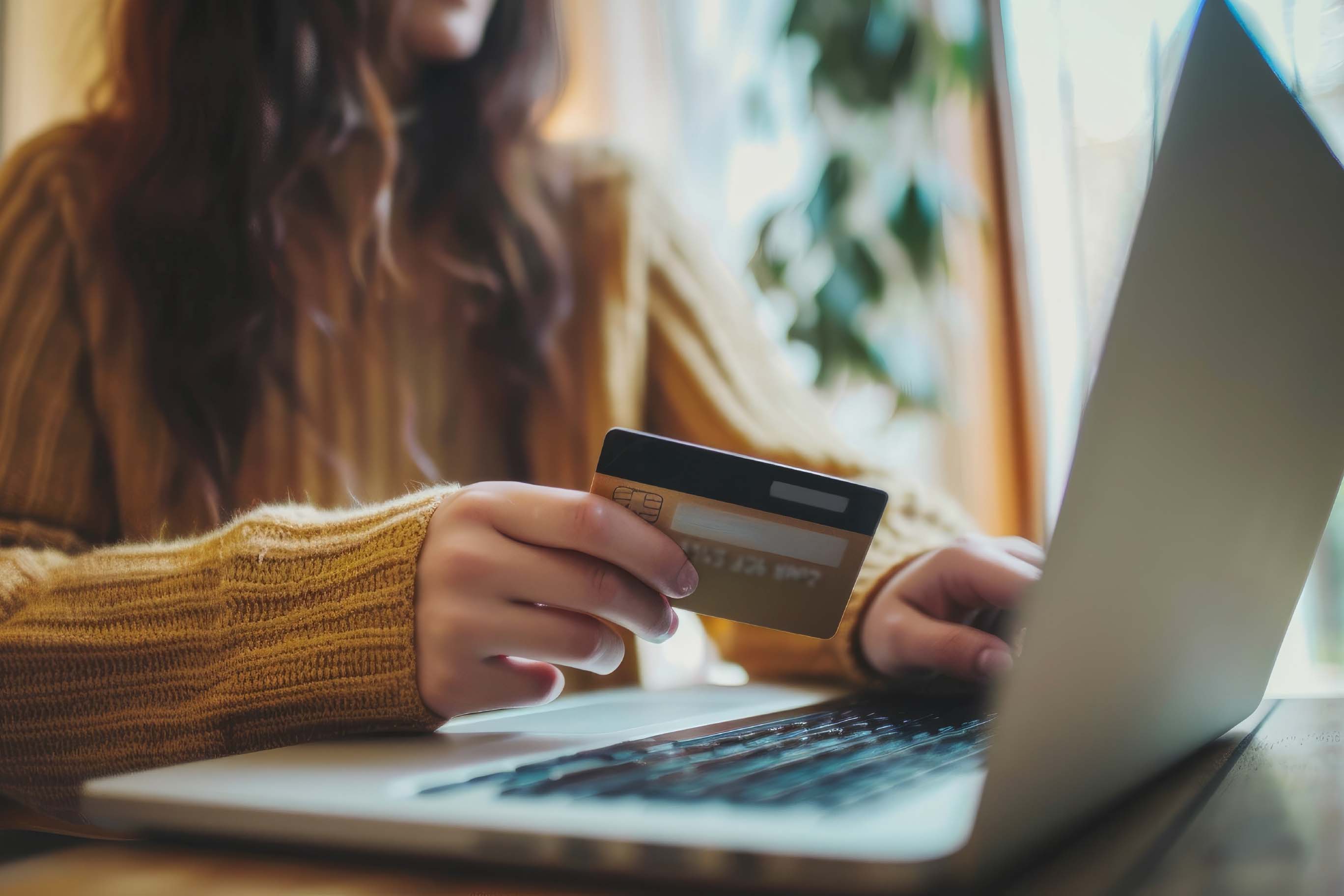 Close-up view of a person wearing a sweater, holding a gold-colored credit card in one hand while resting the other hand on the keyboard of a laptop, appearing ready to make an online payment.